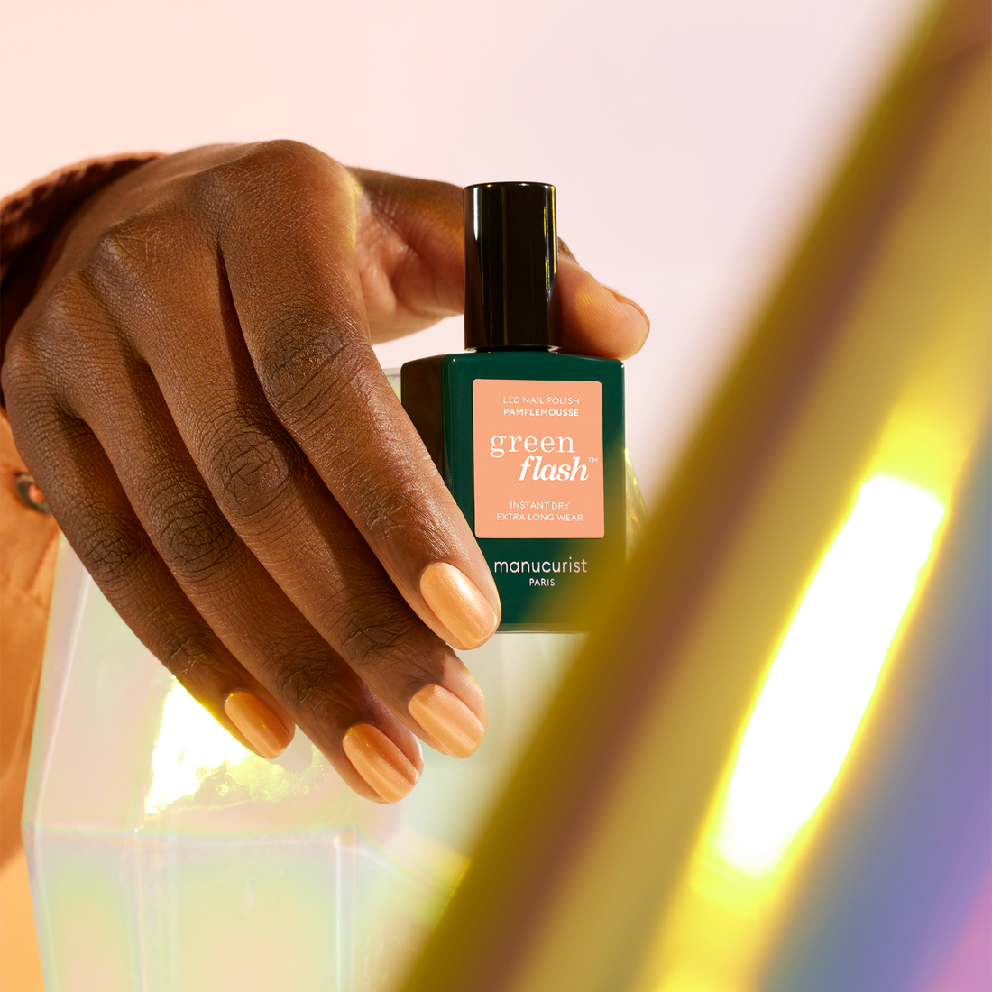 A hand with neatly manicured natural nails holds a bottle of Manucurist Pamplemousse polish against a pastel background with a soft rainbow reflection.