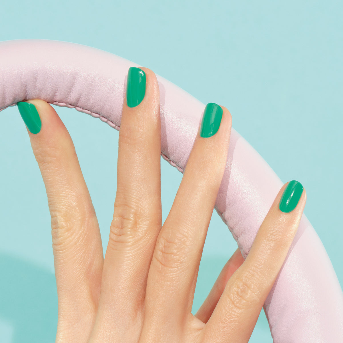Une main au vernis à ongles vert électrique Brazil de Manucurist tient un volant rose clair à texture cuir sur fond bleu clair.