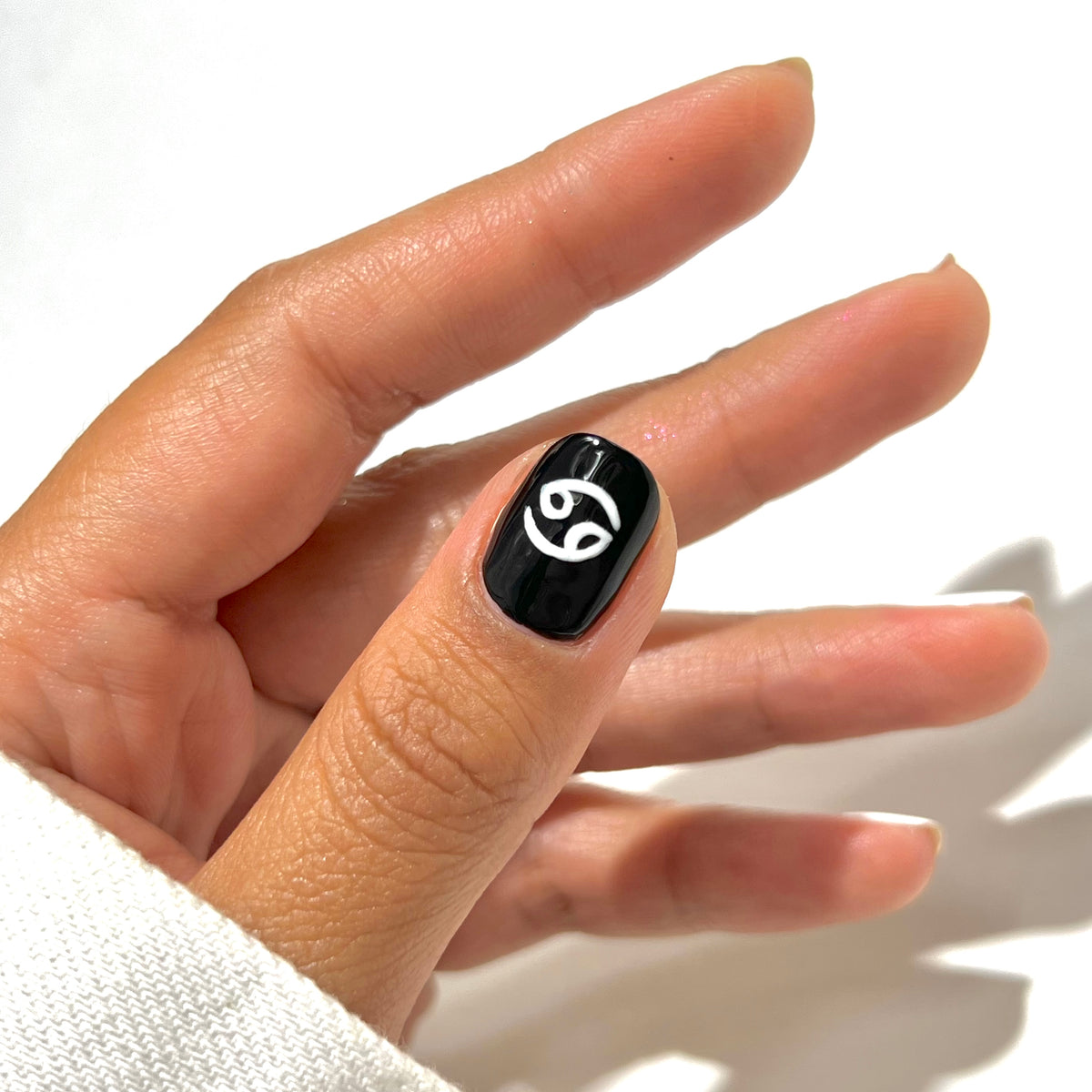 A hand with neatly manicured nails, featuring a black polish and a white Cancer zodiac symbol painted on the thumbnail.