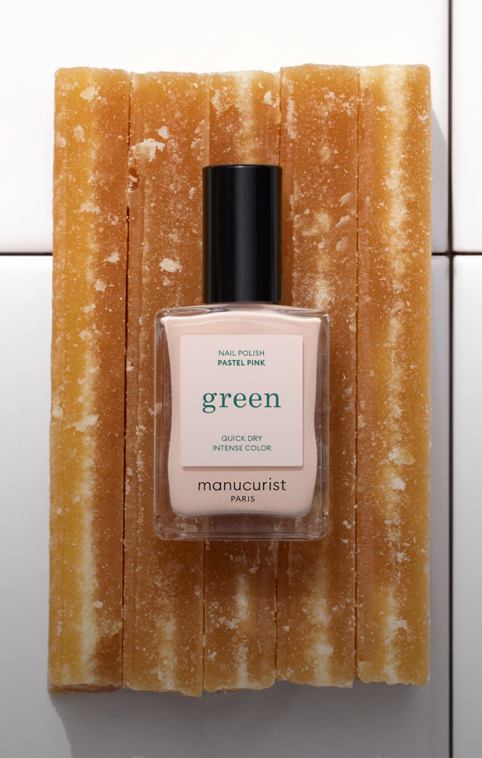 A bottle of Manucurist Paris green pastel pink nail polish is placed upright on top of several stacked caramel-colored rectangular sticks, arranged against a white tiled background.