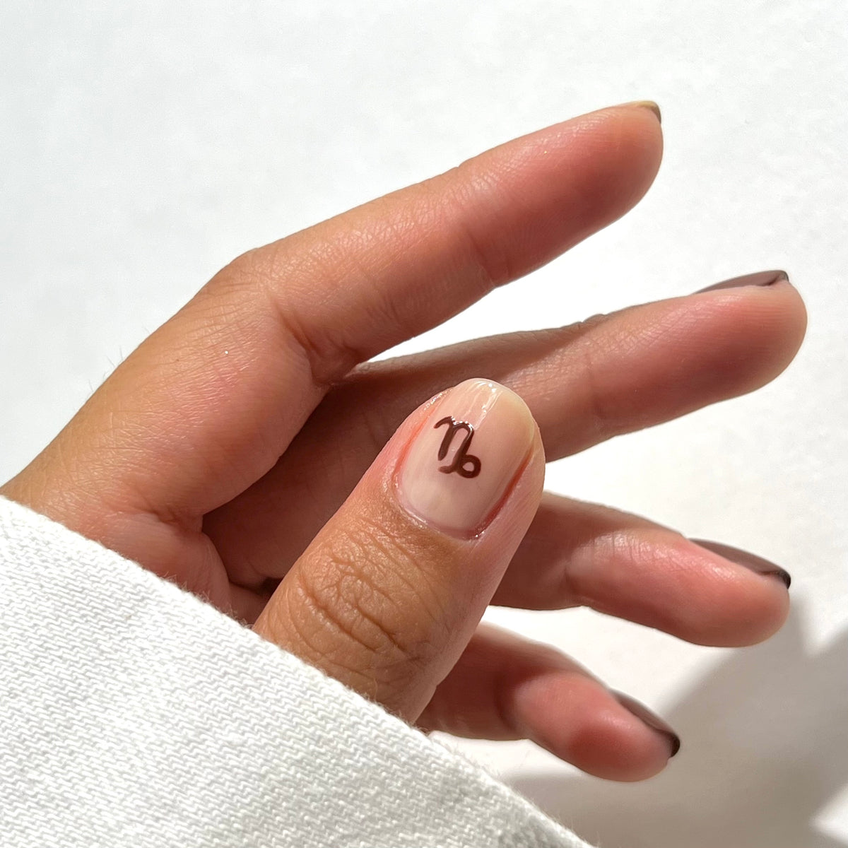 Une main aux ongles soigneusement manucurés, chacun bordé d’une fine ligne marron, avec sur l’ongle du pouce un symbole astrologique du Capricorne marron. La main est partiellement recouverte d’une manche blanche, sur un fond blanc uni.