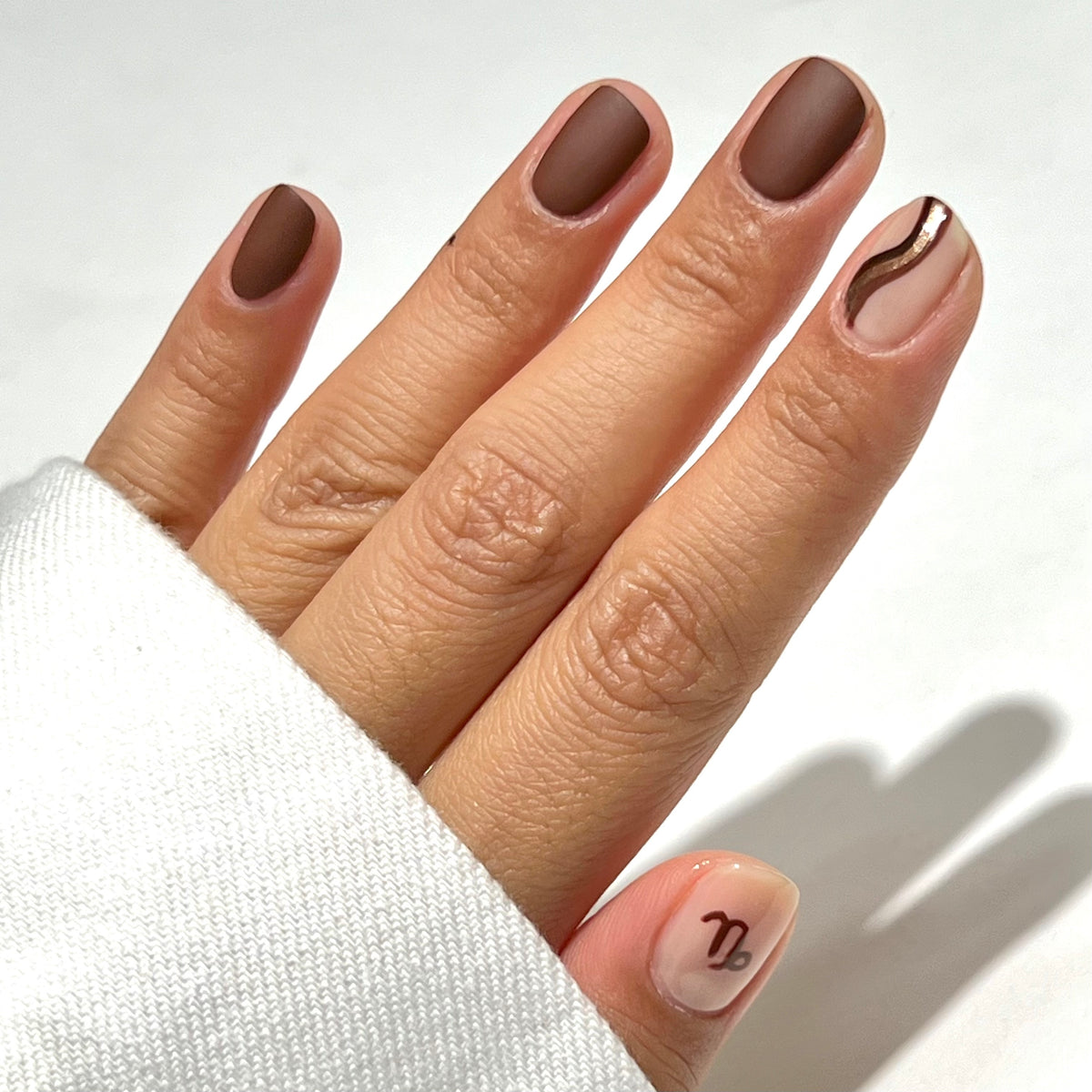 Une main aux ongles courts et manucurés. La plupart des ongles sont vernis d'un marron mat ; l'annulaire présente une base nude ornée d'une ligne ondulée marron, et le pouce est nude avec le symbole du zodiaque du Capricorne en marron. La personne porte une manche blanche.