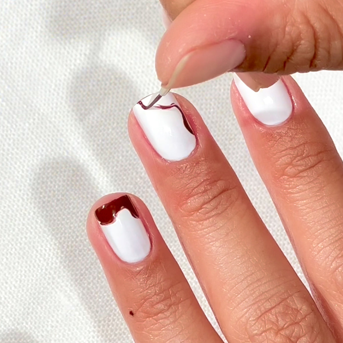 A hand with white-painted nails is shown. A person is using a fine brush to add a wavy brown design to the tip and side of one nail. The background is a white textured surface.