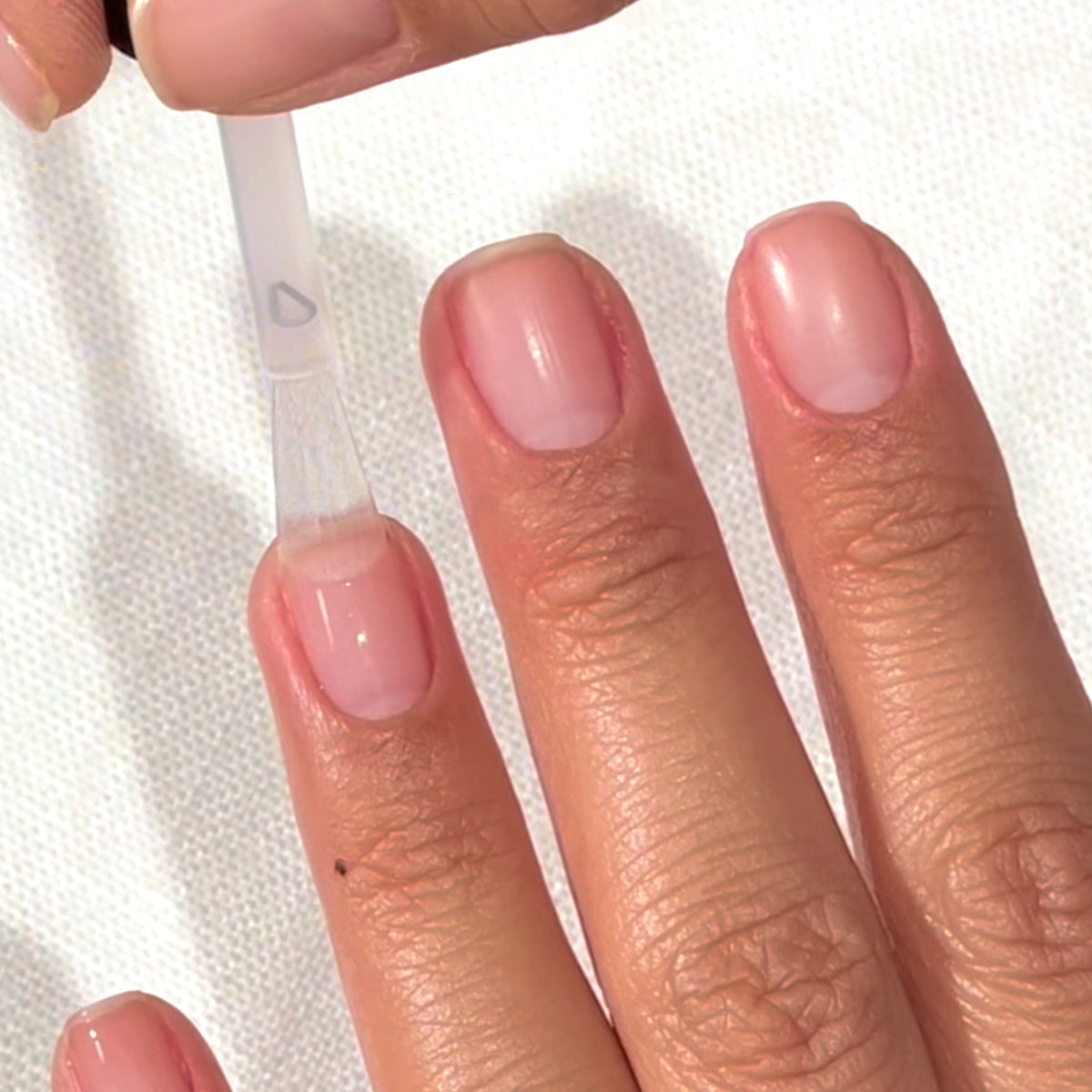 A hand with clean, natural nails is shown as clear nail polish is being applied to the thumbnail using a brush. The background is a white textured surface.
