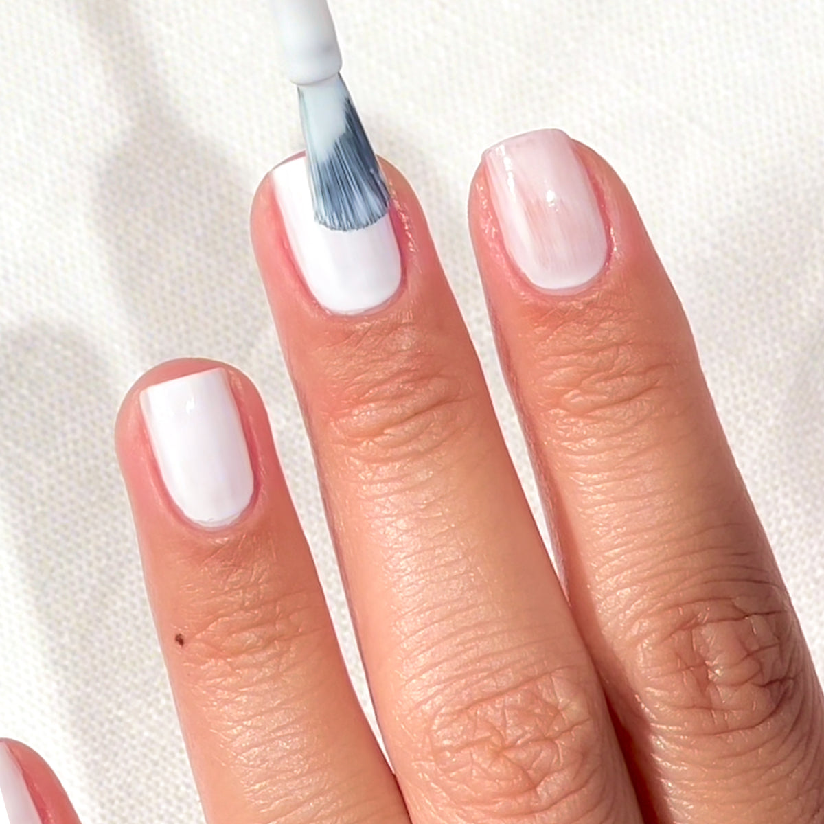 A close-up of a hand with three fingers visible, nails being painted with white nail polish using a brush, against a white textured background.