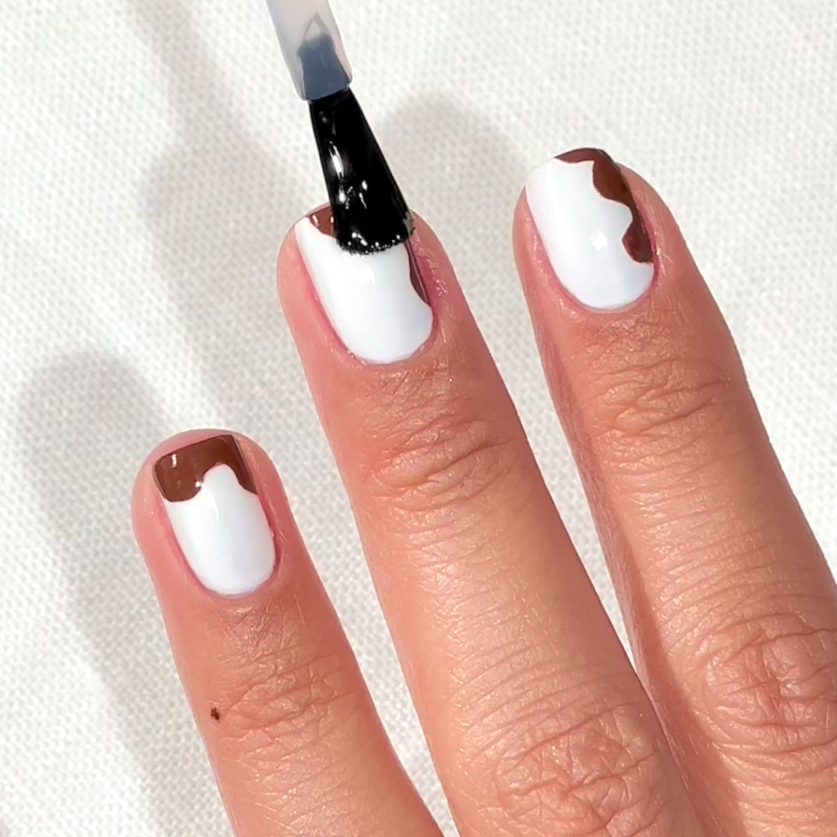 A hand with white nail polish and brown abstract designs on short nails, with a brush applying a clear top coat to one nail. The background is a textured white surface.