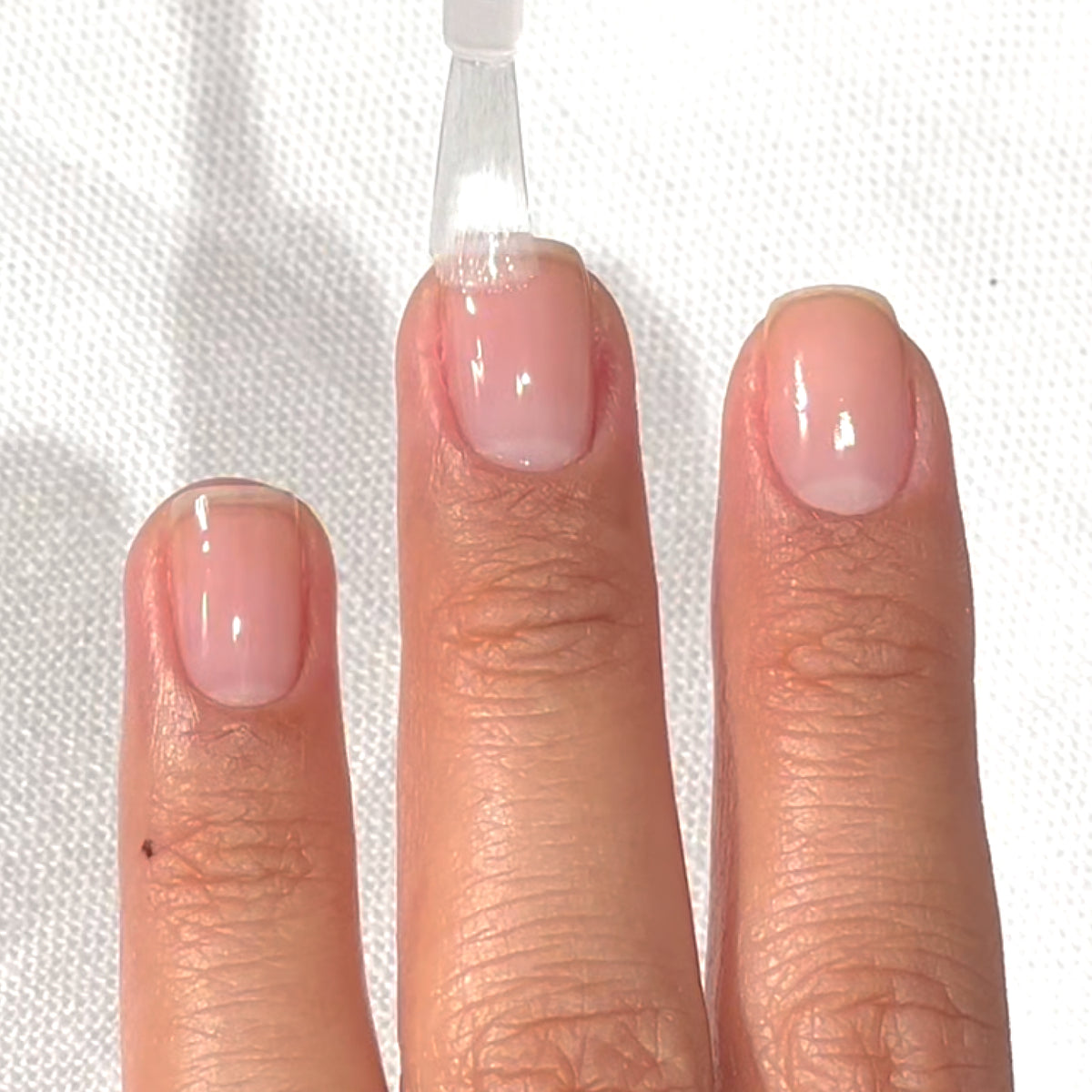 A hand with short, natural nails is shown as a clear nail polish is being applied to the middle fingernail over a light-colored surface.