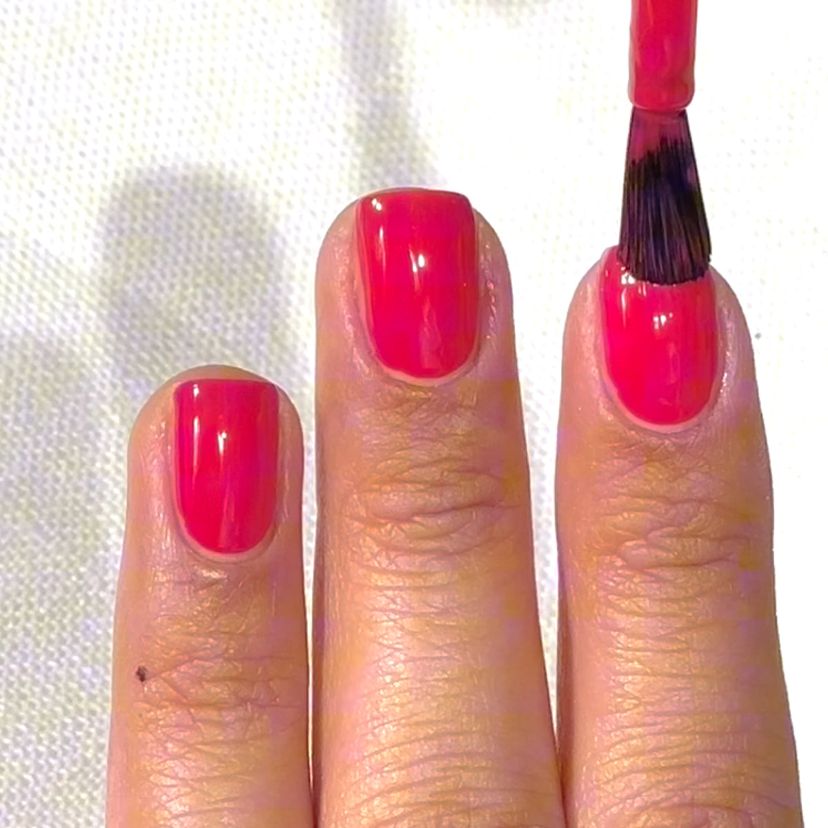 A hand with short nails is having bright pink nail polish applied to the middle fingernail using a brush. The other nails are already painted.