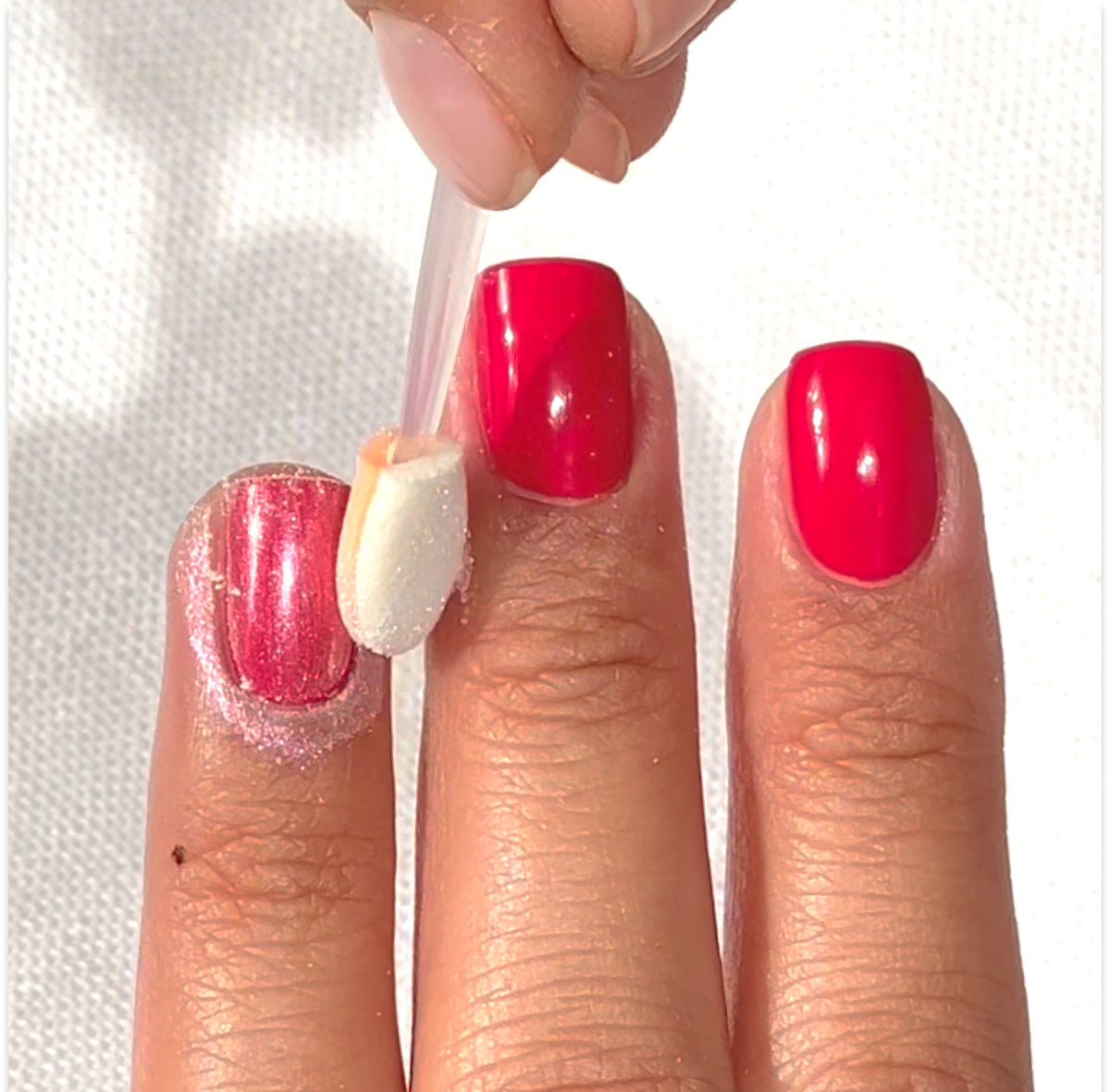 A hand with bright pink nail polish is shown. One finger is being brushed with a sponge applicator, likely applying shimmer or powder to the nail, while the other nails remain untouched.