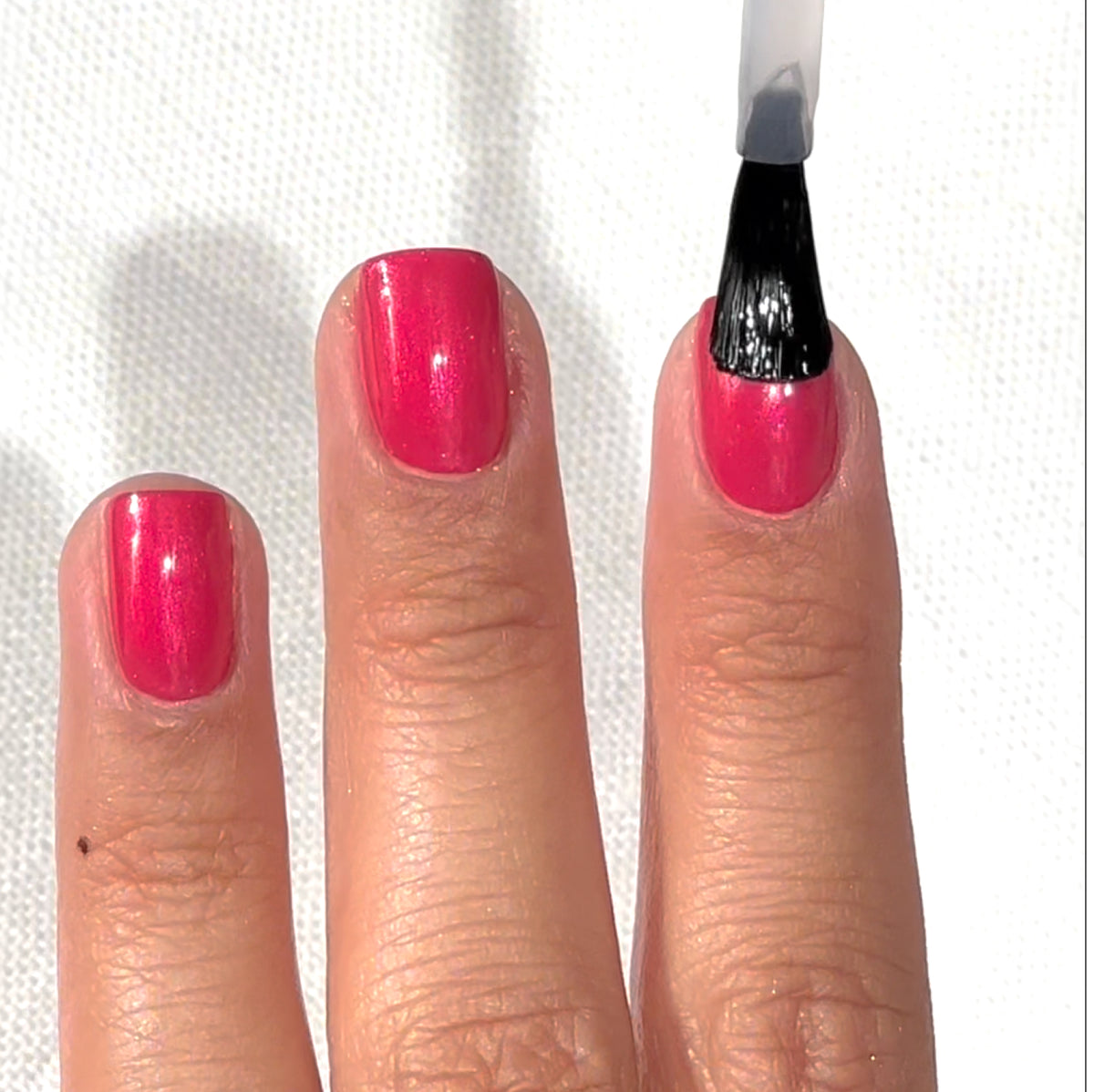 A hand with pink nail polish is shown, while a brush applies clear top coat to the middle fingernail. The background is a white textured surface.