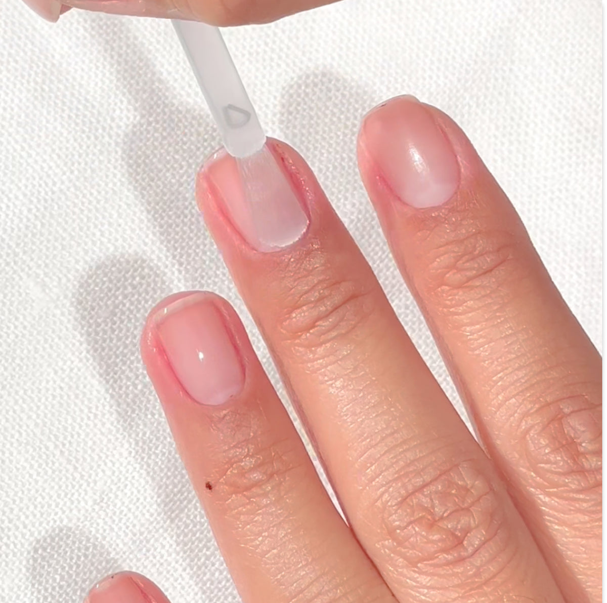 Gros plan sur une main aux ongles courts et naturels, tandis qu’un vernis transparent est appliqué au pinceau sur l’ongle du majeur. Le fond est une surface blanche texturée.