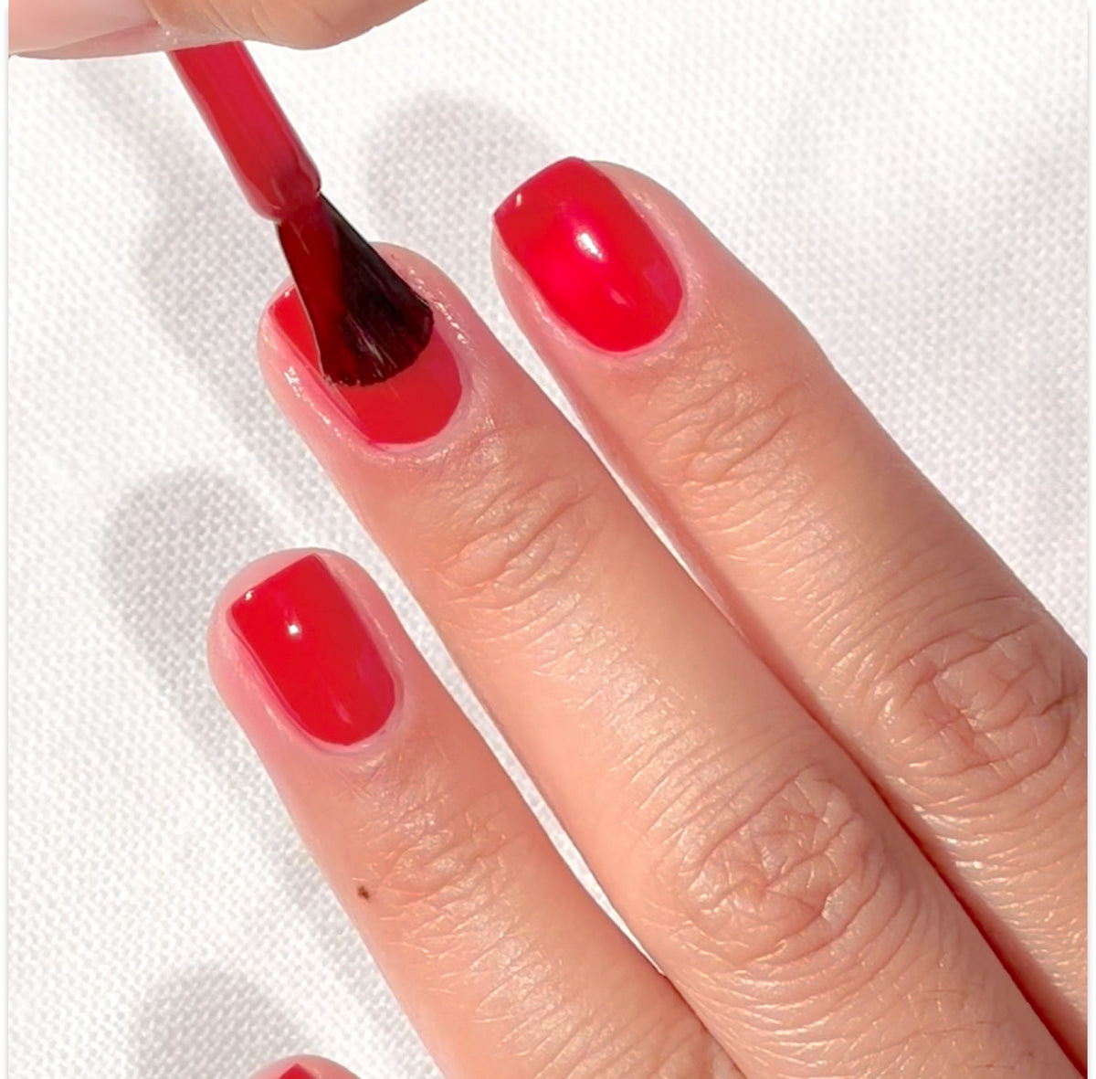 Une main aux ongles soigneusement coupés reçoit l’application d’un vernis à ongles rouge vif à l’aide d’un pinceau, sur un fond blanc texturé.