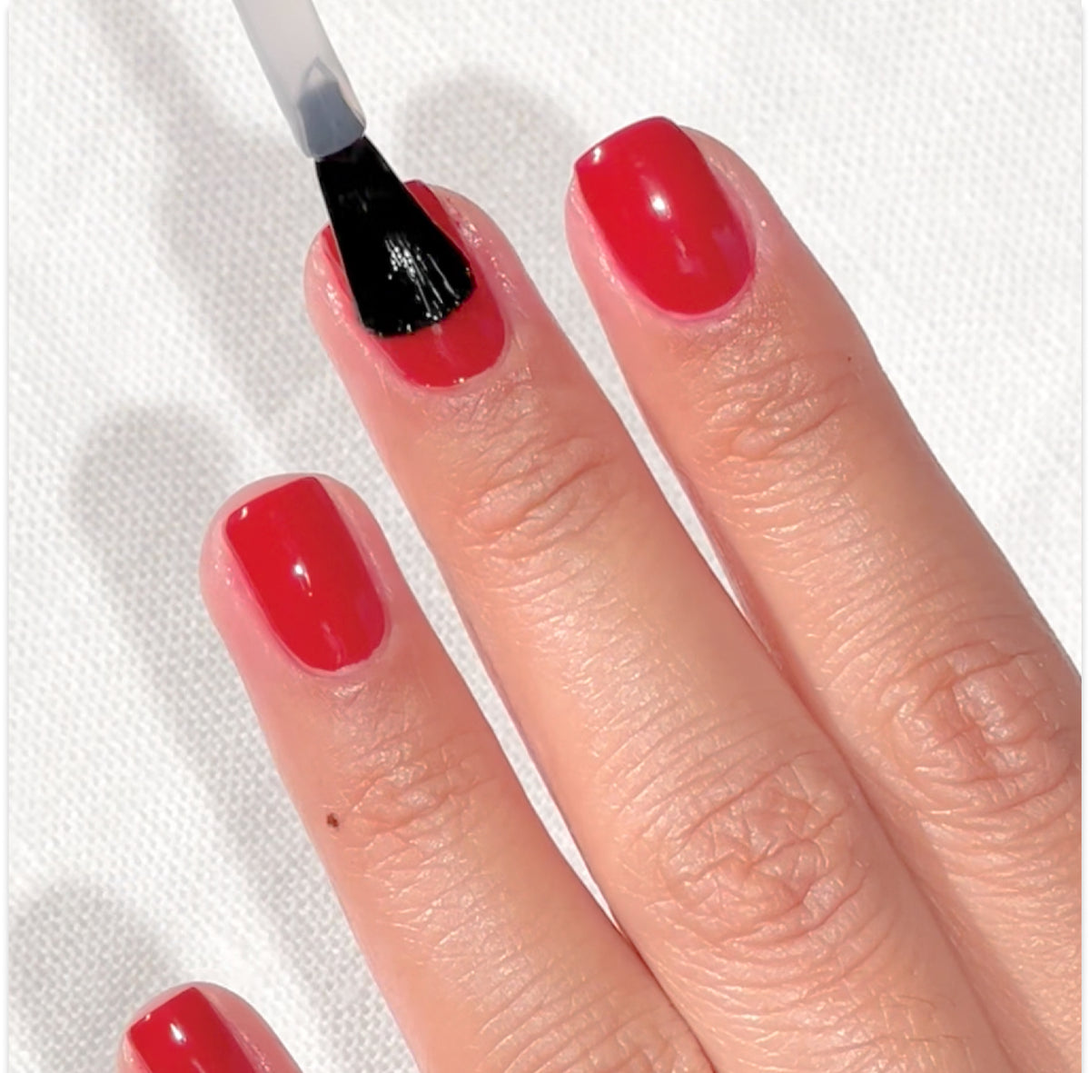 Une main aux ongles peints en rouge est montrée, et un pinceau applique un top coat transparent sur l'ongle du majeur. L'arrière-plan est une surface blanche texturée.