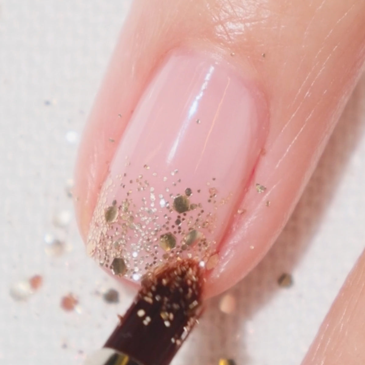 Gros plan d'un ongle verni d'un rose translucide et de paillettes dorées vers le bout, appliqué à l'aide d'un petit pinceau. Les paillettes sont parsemées sur l'ongle et la peau autour.
