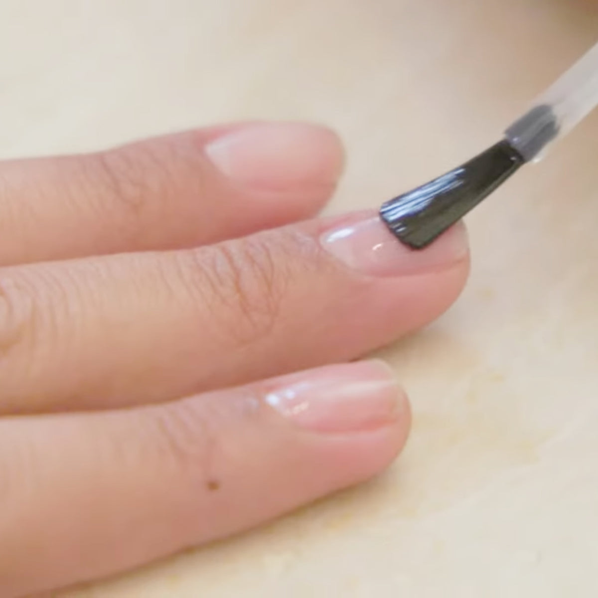 A close-up of a hand with natural nails as a clear nail polish is being applied to the fingernail with a brush.