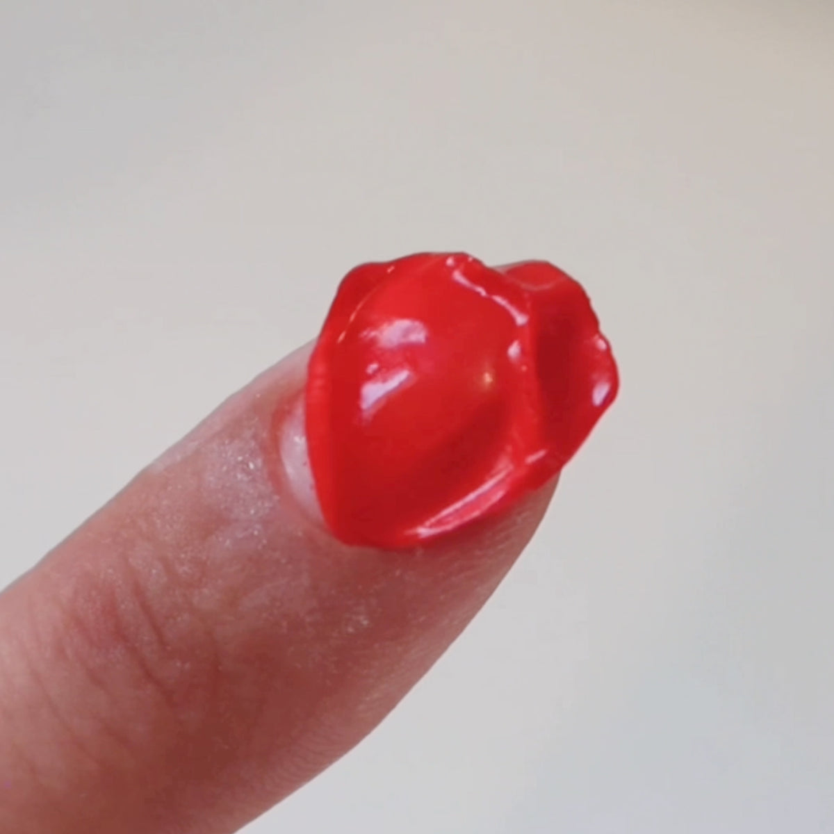 A finger with a shiny, bright red blob of substance on the tip, shaped somewhat like a small heart, against a plain light background.