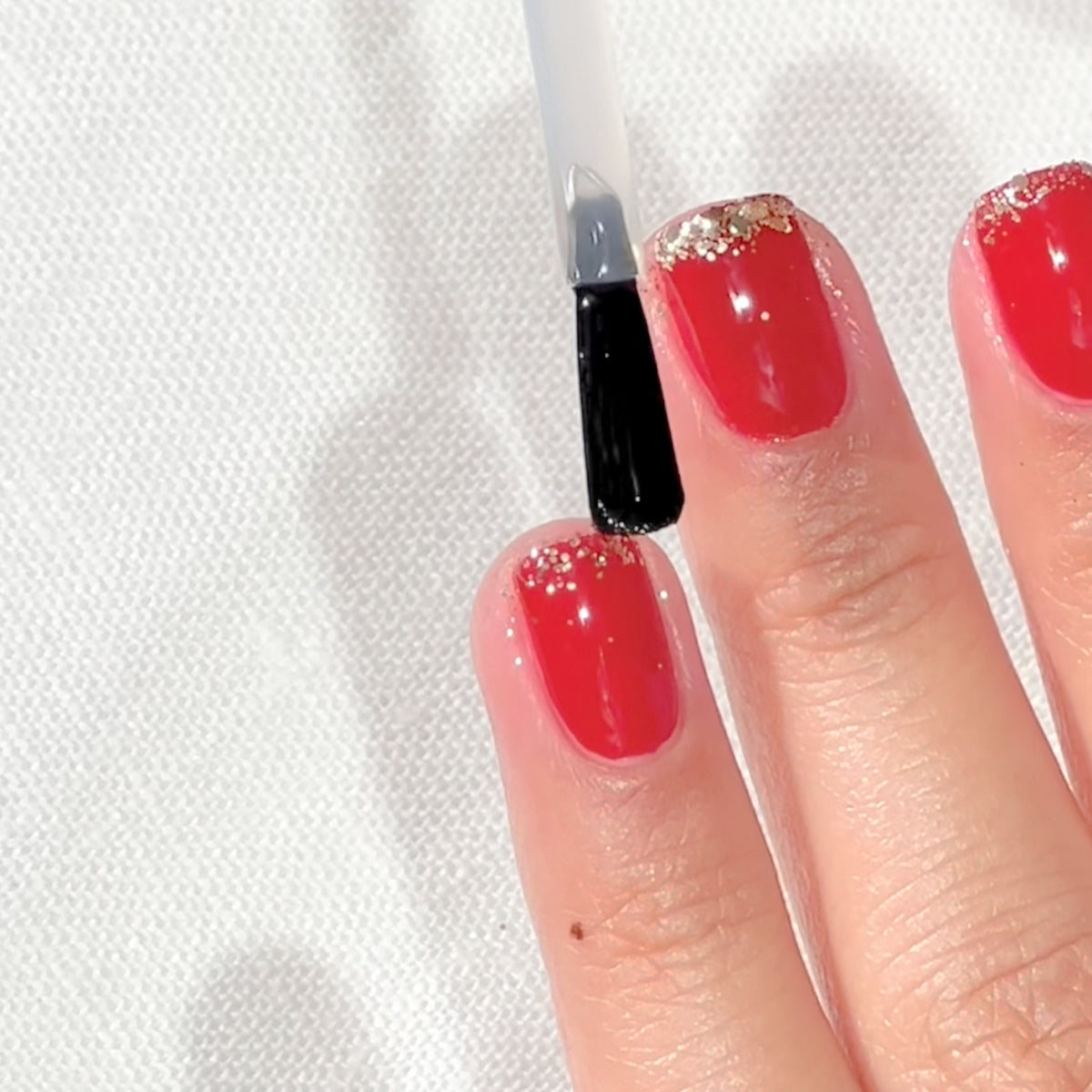 Une main avec un vernis à ongles rouge et des pointes à paillettes dorées reçoit l'application d'un top coat transparent sur l'ongle du majeur à l'aide d'un pinceau.
