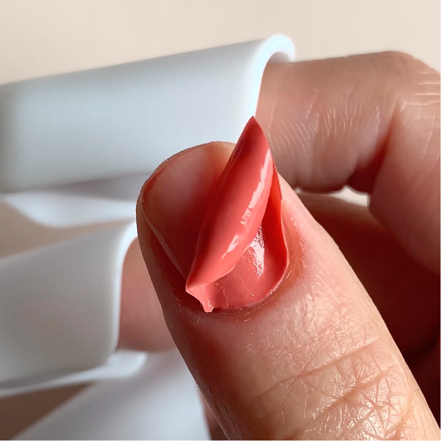 A close-up of a thumb with coral-colored nail polish being peeled off in one piece, held between fingers with a white object in the background.