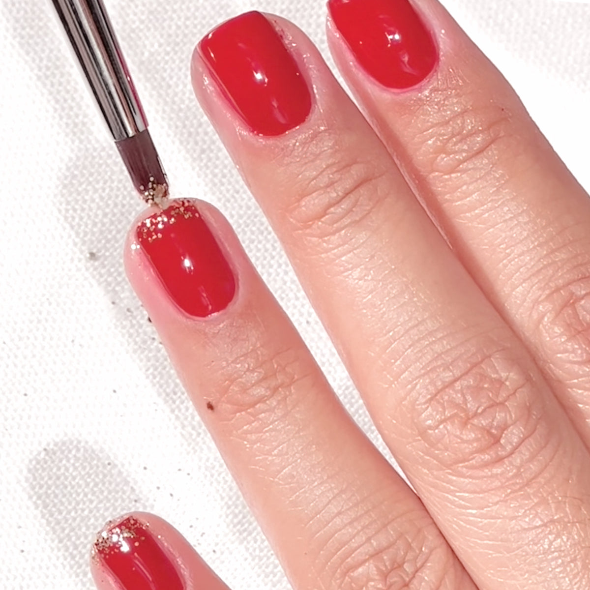 Une main aux ongles vernis de rouge est décorée de paillettes dorées à la base des ongles à l’aide d’un petit pinceau.