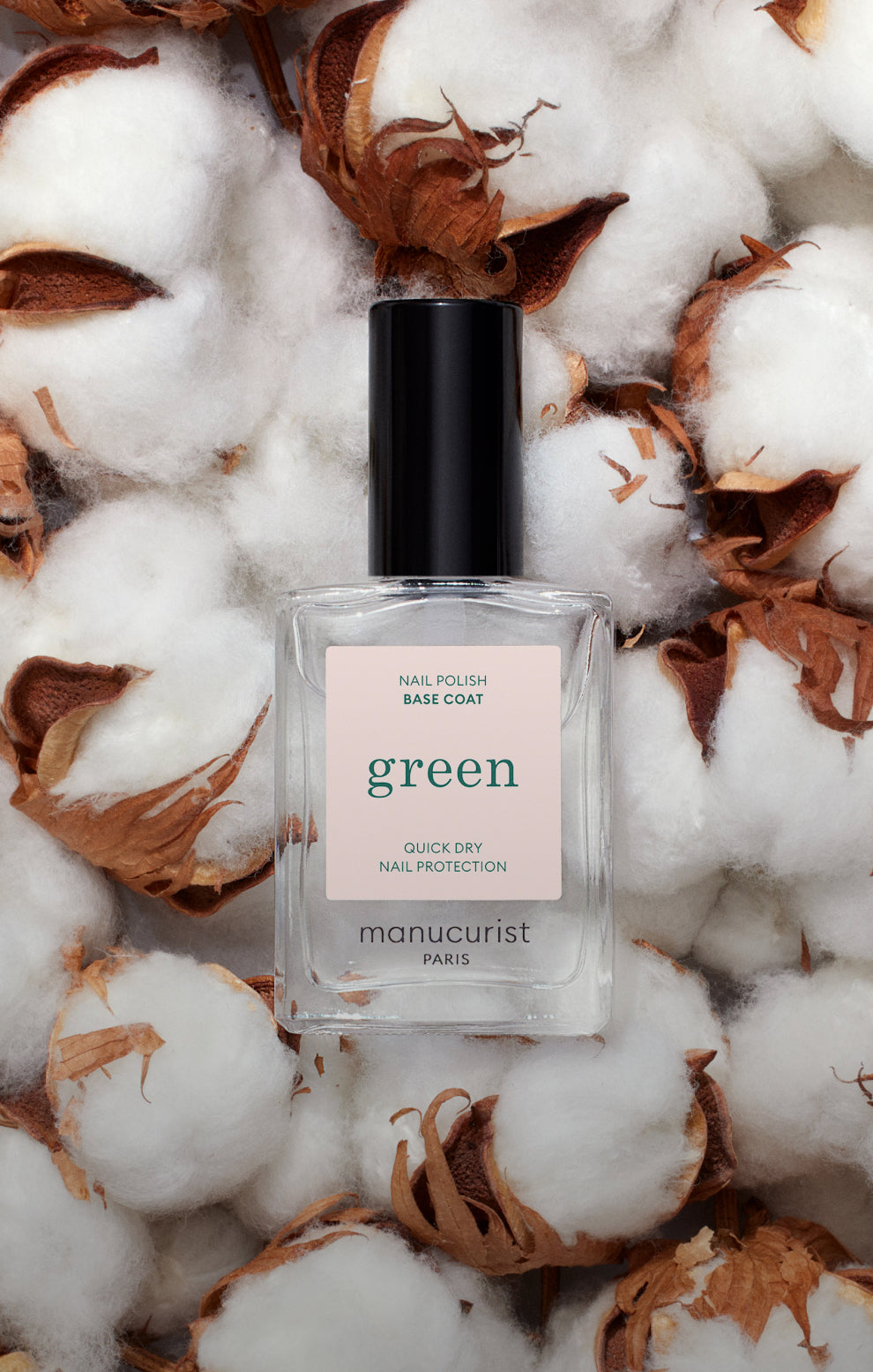 A clear bottle of Manucurist Paris green nail polish base coat with a black cap is placed on a bed of fluffy cotton bolls.