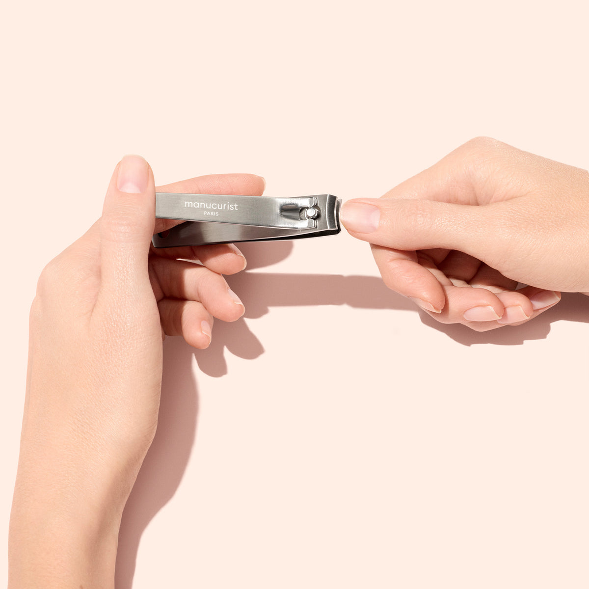 A person uses a silver nail clipper to trim the fingernails on their left hand against a pale pink background.