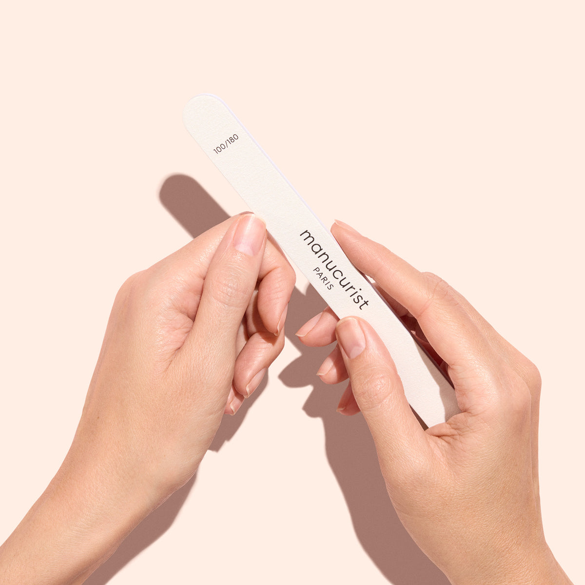 Two hands holding a white nail file labeled manucurist Paris against a light peach background.