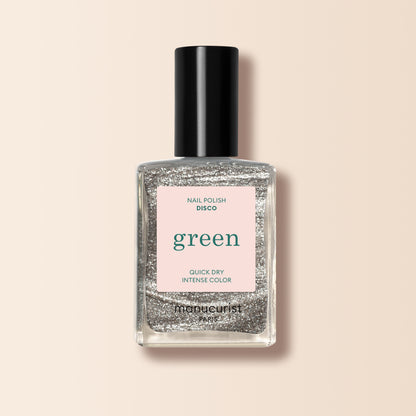 A clear glass bottle of Manucurist Disco silver glitter nail polish from the Green™ range, labeled green and quick dry intense color, stands upright against a light beige background.