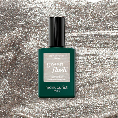 A Manucurist Disco gel polish bottle sits on a shimmering silver background. The bottle is dark green with a black cap and features a sparkly silver label.