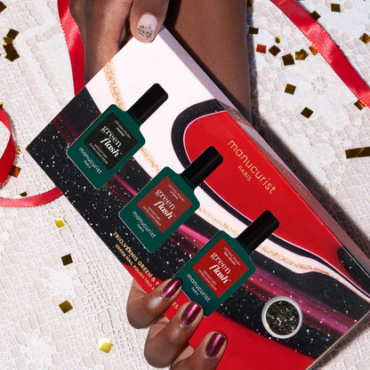 A glittery-manicured hand holds the Manucurist-test Midnight Glam Trio + Glitter Set gift box, displaying three green polishes with biodegradable glitter, on a lace table decorated with gold confetti.