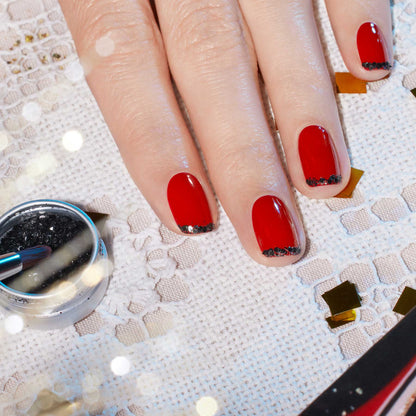 Une main aux ongles courts et arrondis, vernis rouge vif et ornés de Manucurist Black Diamond Biodegradable Glitter, repose sur de la dentelle. À proximité se trouvent un petit pot de paillettes et un pinceau de nail art.