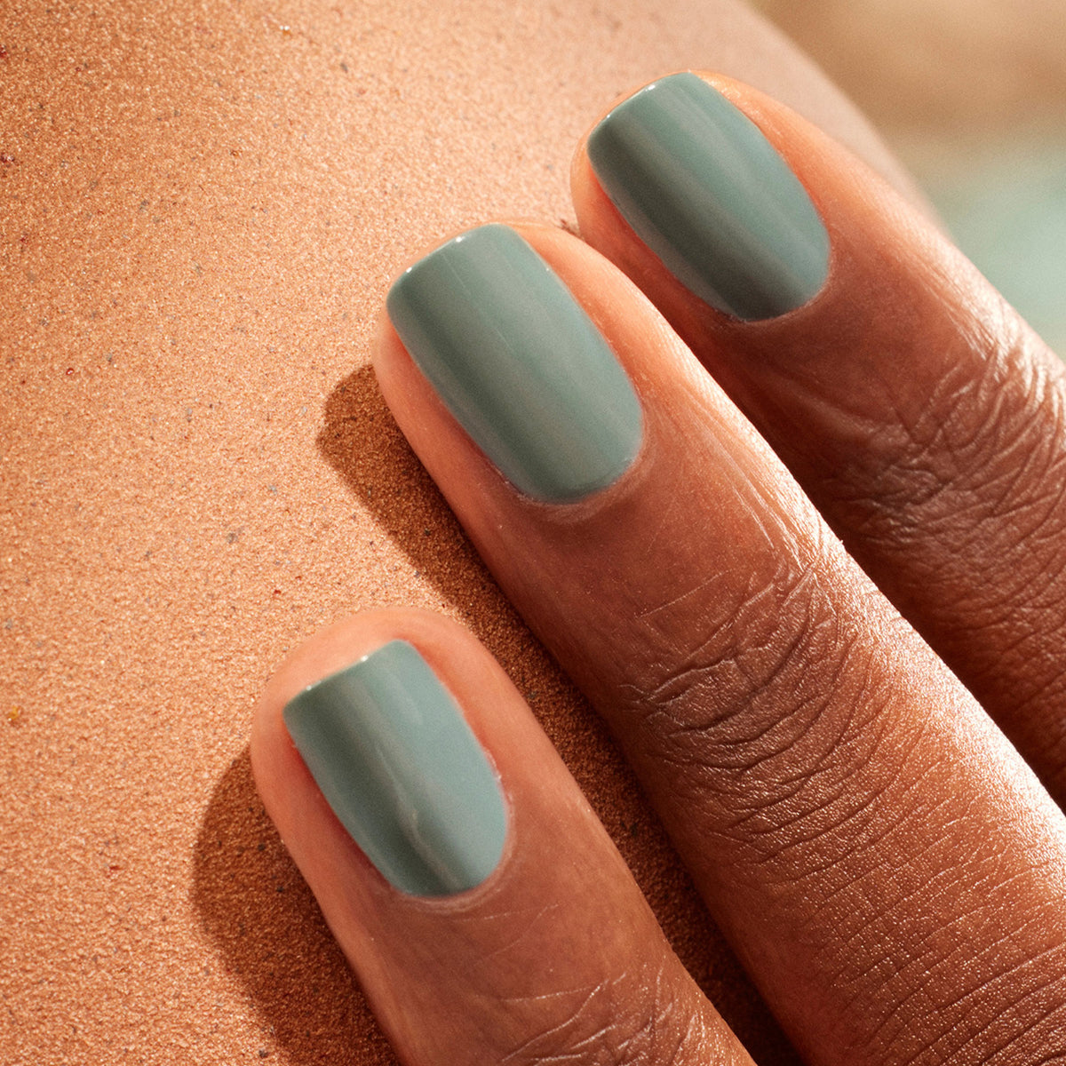 Gros plan d’une main à la peau foncée aux ongles soigneusement manucurés, vernis avec Sage de Manucurist, une teinte vert tendre, posée sur une surface texturée.
