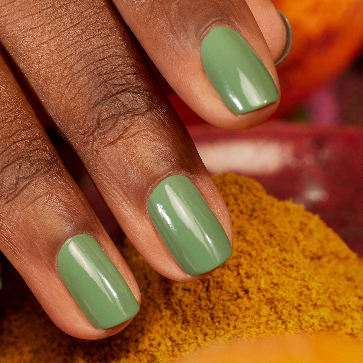 Gros plan sur une main à la peau brun moyen, aux ongles vernis dans la teinte brillante "Artichoke" de Manucurist. Les tons jaune chaud et orange en arrière-plan évoquent l’atmosphère d’un jardin marocain.