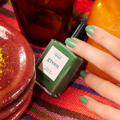 Une main aux ongles vernis de vert tient un flacon de vernis à ongles Manucurist étiqueté Artichoke, posé sur un tissu tissé vibrant et entouré de céramiques rouges et orange rappelant l&