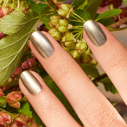Gros plan sur une main arborant des ongles argentés brillants et froids Manucurist Metallic tenant une grappe de baies vertes et rougeâtres, avec des feuilles en arrière-plan.