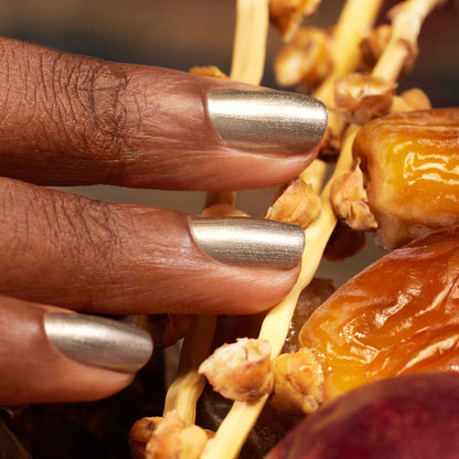 Des doigts vernis avec le vernis Manucurist Metallic dans une teinte argentée aux sous-tons froids touchent une grappe de dattes séchées sur leur branche.