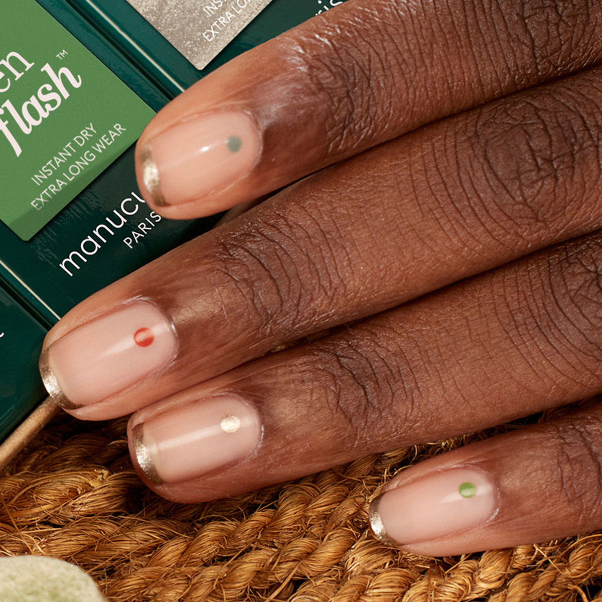A close-up of a hand with a minimalist manicure using Manucurist’s Spice Lover from the Autumn 2023 collection, featuring clear polish, metallic gold French tips, and a colored dot. The hand rests on textured brown near Green Flash packaging.