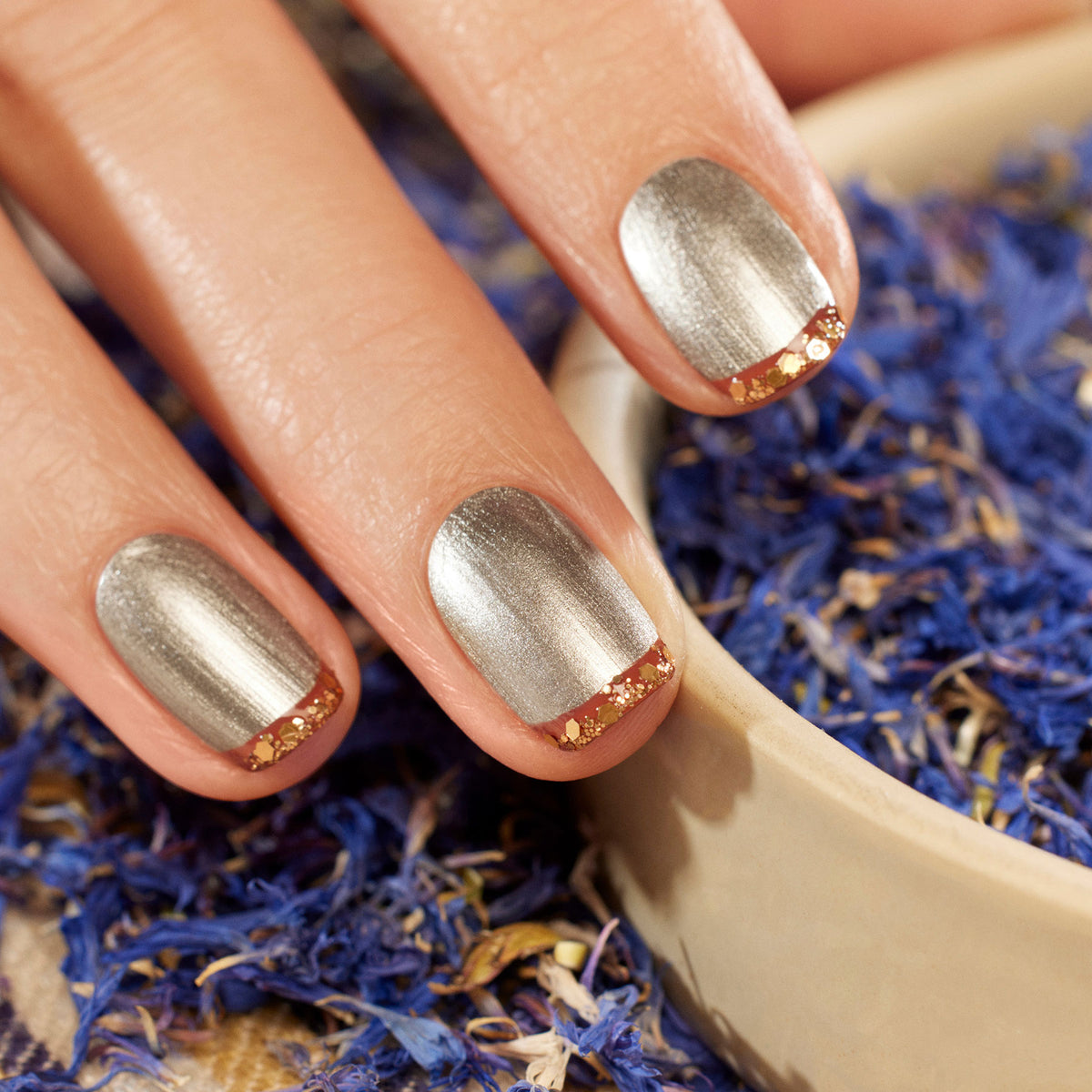 Une main avec du vernis à ongles argent métallisé et des pointes de paillettes biodégradables Copper de Manucurist repose sur le bord d’un bol rempli de pétales bleus et violets séchés.