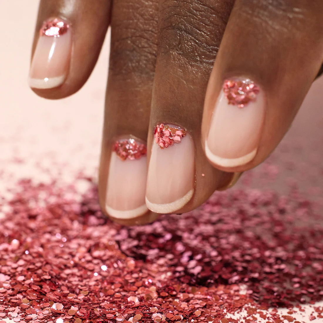 A hand with light pink French manicure nails, each decorated with pink glitter near the cuticles, is shown above a surface covered in scattered pink glitter.
