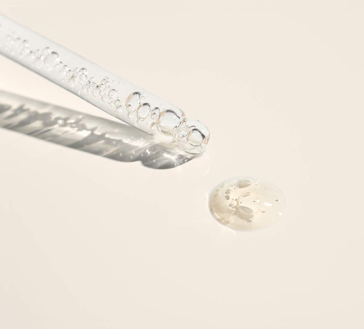 A clear glass dropper with bubbles inside lies on a light beige surface next to a drop of transparent liquid, casting a soft shadow.