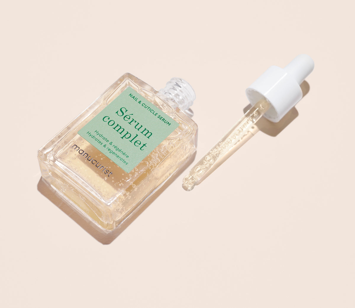 Un flacon en verre transparent du sérum ongles et cuticules Sérum Complet à étiquette vert pâle est posé à côté de son bouchon pipette, sur un fond beige clair. La pipette contient une petite quantité de sérum.