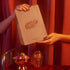 Two hands with red painted nails hold a beige Small Gift Bag labeled Manucurist in front of red curtains. The nail care gift shines, and below it, a glass ornament and bowl of baubles sparkle on the red velvet surface.