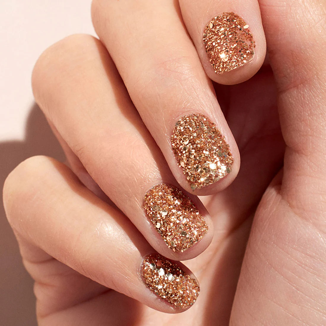 A close-up of a hand with short, neatly shaped nails painted with gold glitter nail polish. The fingers are gently curled, and the background is a soft, light color.