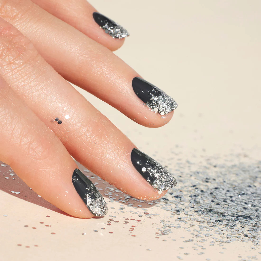 A hand with black nail polish featuring silver glitter ombré tips rests on a light surface, surrounded by scattered silver glitter.