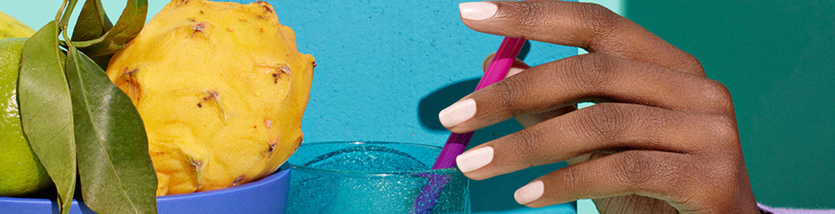 A hand with light pink nail polish holds a pink straw in a blue glass, next to a bowl with a yellow dragon fruit and green citrus fruits against a colorful blue background.
