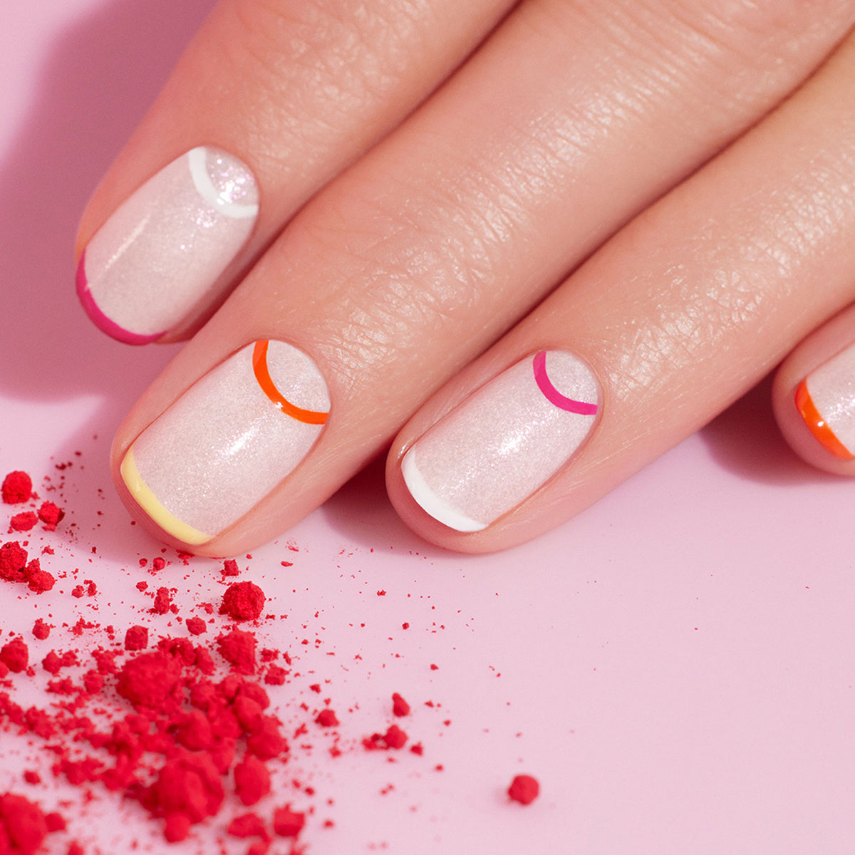 A close-up of a hand with shimmery pale pink “Smile!” nails by Manucurist, featuring colorful minimalist half-moon summer nail art in orange, red, pink, yellow, and white. Crushed pink powder is scattered nearby on a pink surface.
