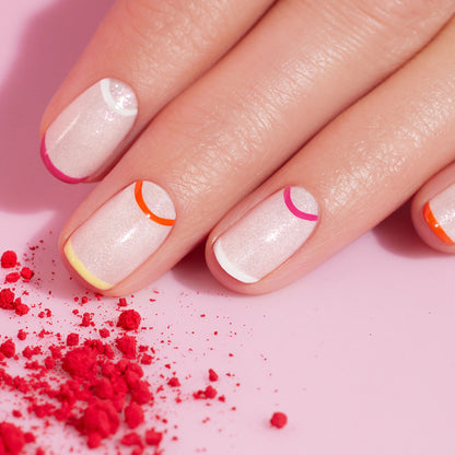 A close-up of a hand with shimmery pale pink “Smile!” nails by Manucurist, featuring colorful minimalist half-moon summer nail art in orange, red, pink, yellow, and white. Crushed pink powder is scattered nearby on a pink surface.