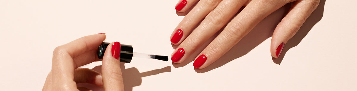 A hand applies clear top coat nail polish to the fingernails of another hand, which has already been painted with glossy red nail polish, against a light-colored background.
