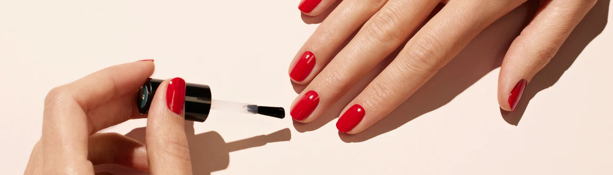 A close-up of a person applying clear nail polish to fingernails painted with bright red nail polish on a light background.