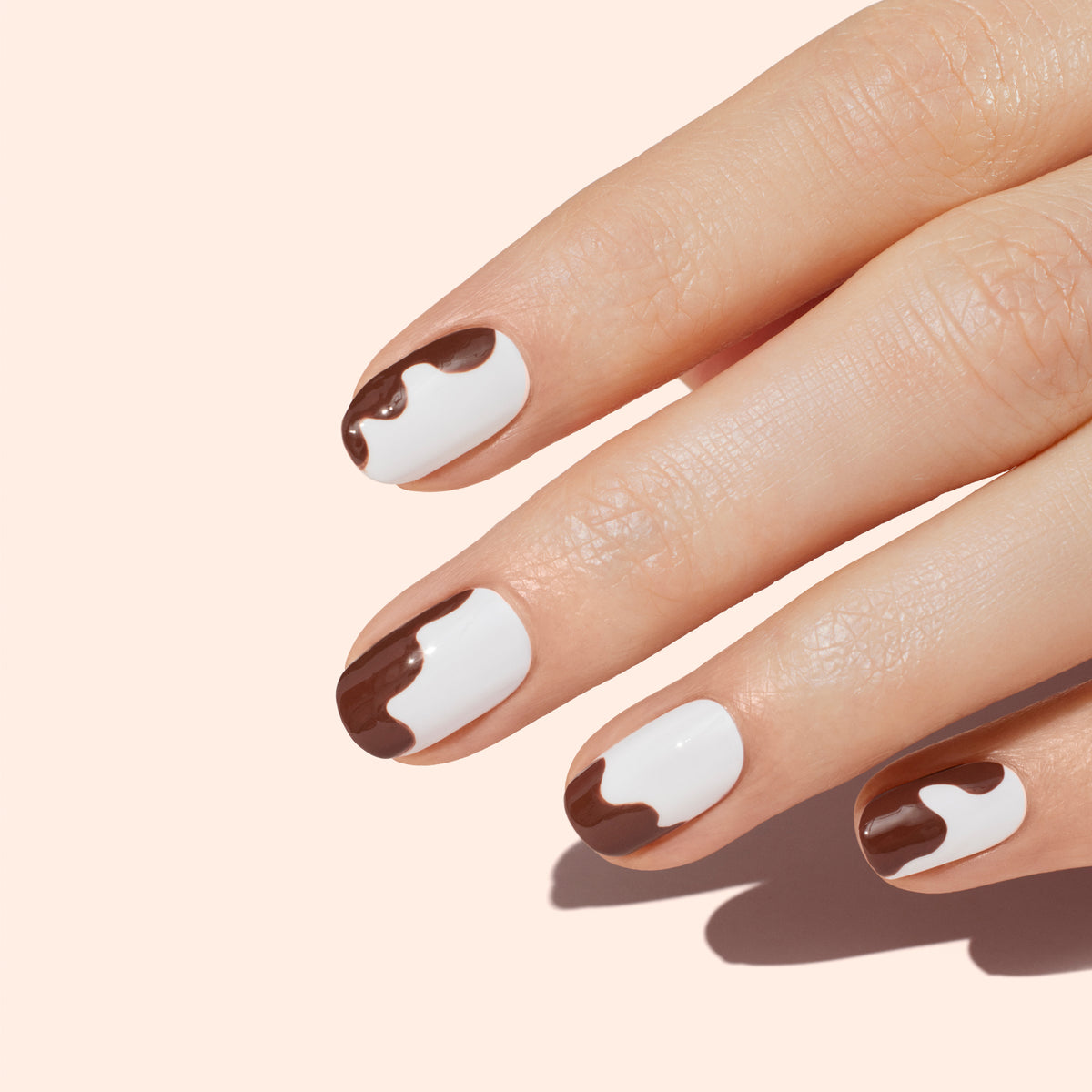 A hand with neatly manicured nails features a unique design: white nail polish with brown, drip-like patterns near the tips, against a pale peach background.