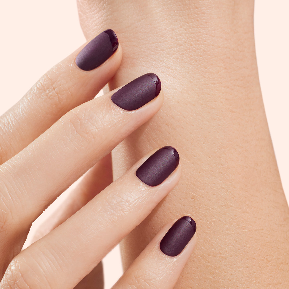 A hand with neatly manicured nails painted in a matte dark purple shade, each nail featuring a glossy dark purple French tip, touches the wrist of the other hand.